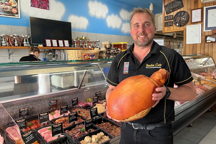 A man wearing a black polo shirt holding a large ham in front of a butcher's cabinet