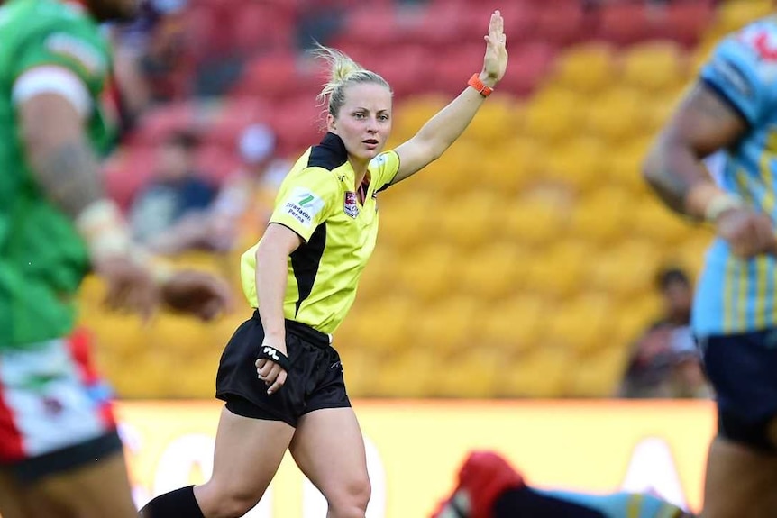 A female rugby league referee runs and raises her left arm as she officiates a match.