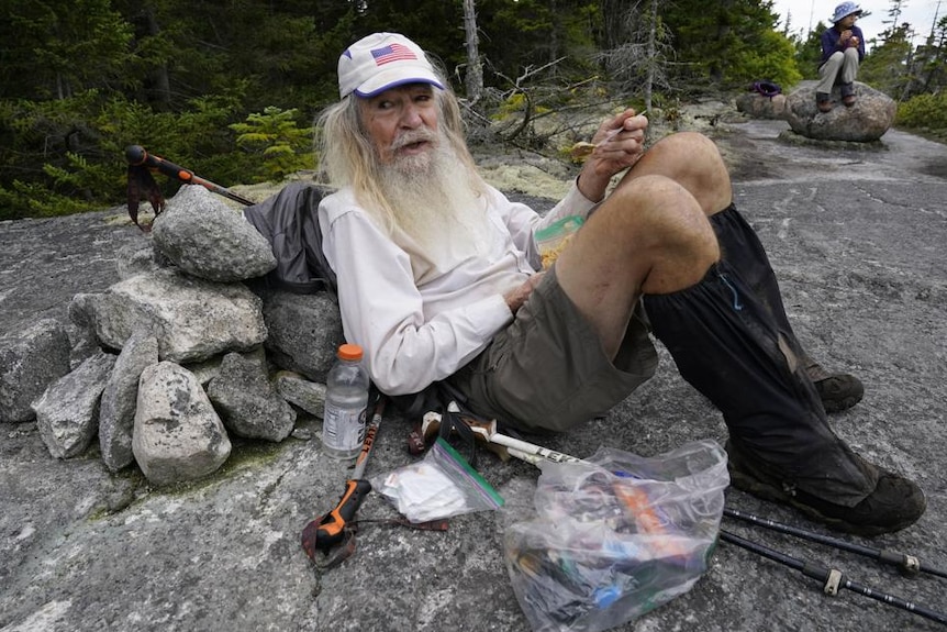 A man in a long sleeved white top and knee length boots resting against rocks while hiking the Appalachian Trail