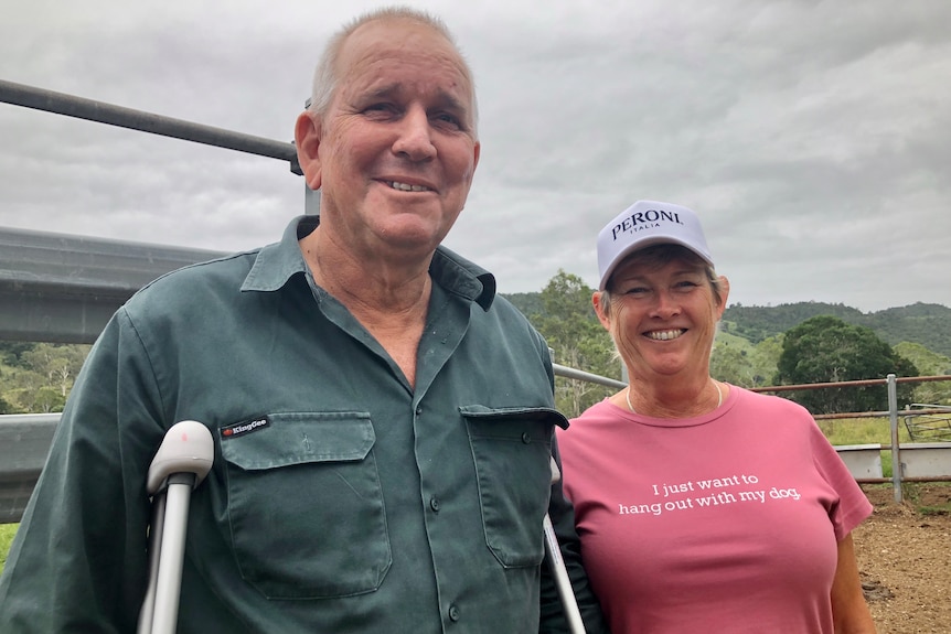 A man and woman smile at the camera with cattle yard railings and mountains behind them.