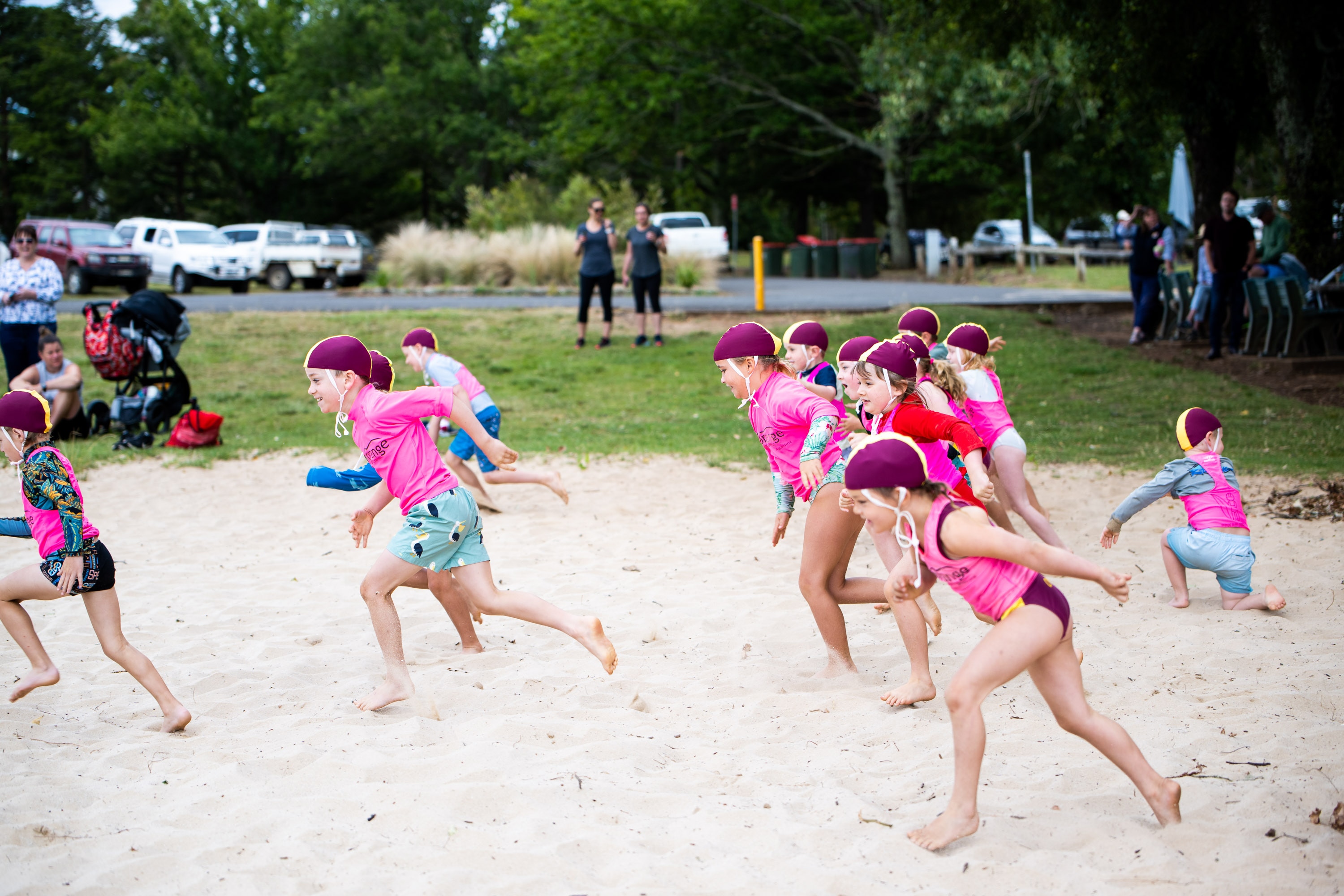A number of kids in swimmers, pink vests and swimming caps run across sand.