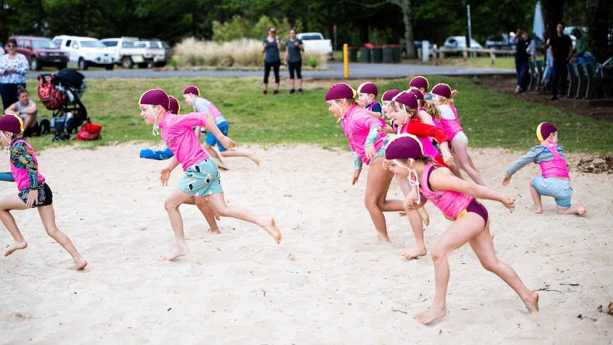 A number of kids in swimmers, pink vests and swimming caps run across sand.