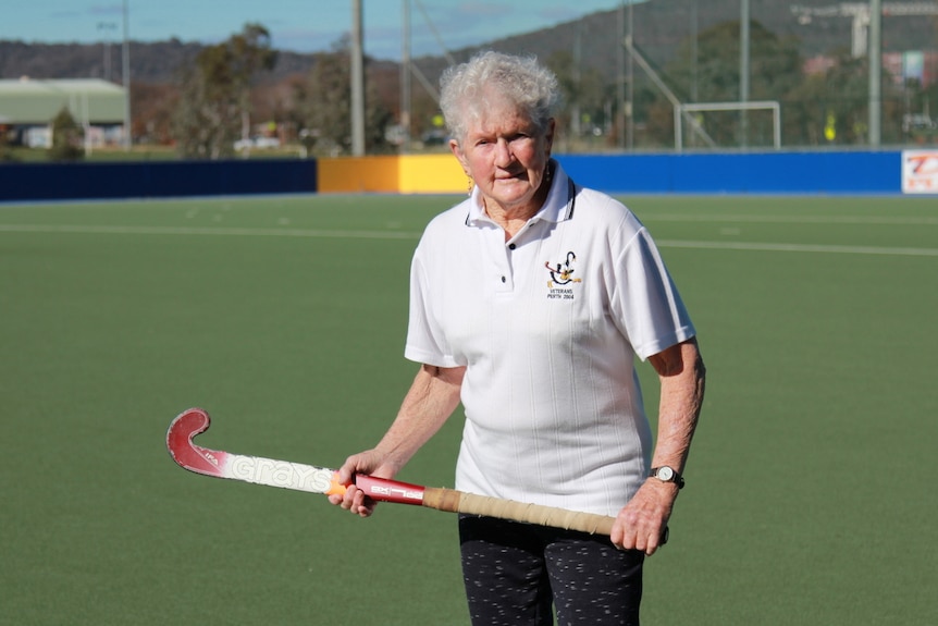 Elderly woman stands on hockey field holding her hockey stick in front of her.