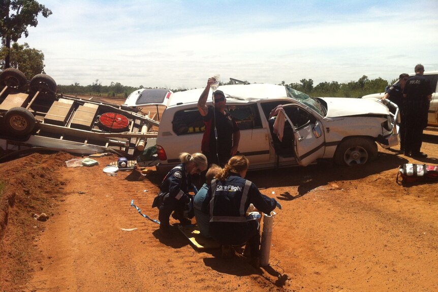 A crashed car and boat on a remote dirt road, with paramedics responding.