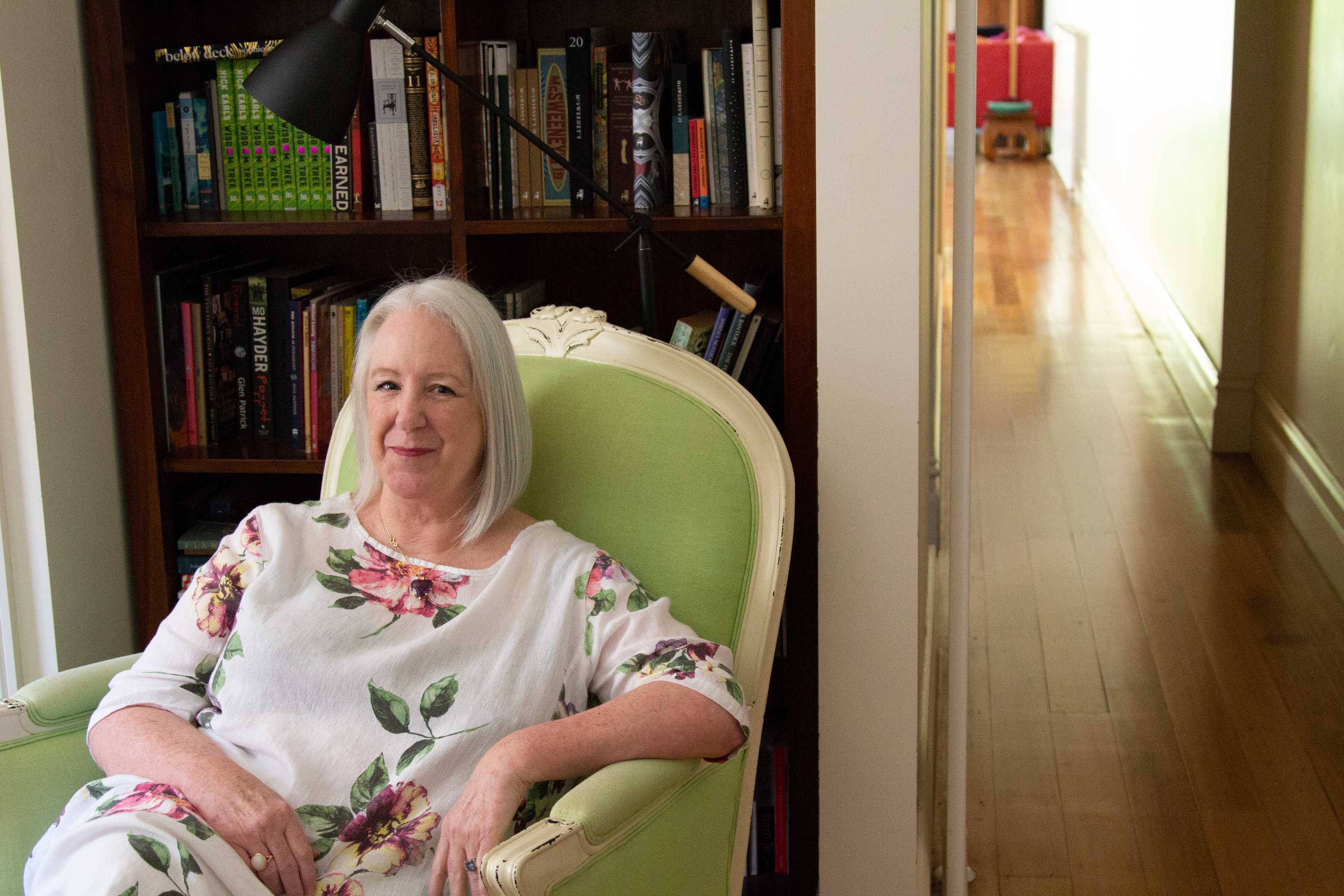 A woman in floral print dress smiles, sitting in a green armchair. Behind her is bookshelf and to the side a hallway stretches.
