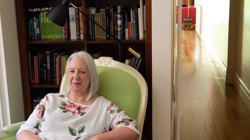 A woman in floral print dress smiles, sitting in a green armchair. Behind her is bookshelf and to the side a hallway stretches.