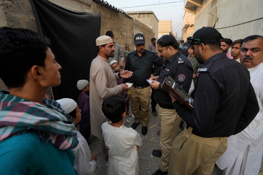 A Police officer checks documents of a resident during a search, while men and boys gather around.