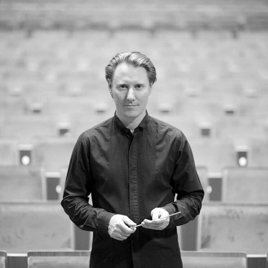 A black and white photo of conductor Benjamin Northey, in black dress, holding his baton at Melbourne's Hamer Hall.