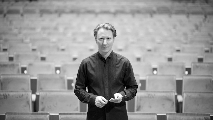 A black and white photo of conductor Benjamin Northey, in black dress, holding his baton at Melbourne's Hamer Hall.