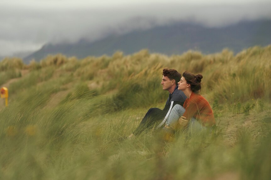 A scene from the TV series Normal People with a young man and woman sitting in some grass on a beach