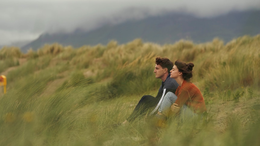 A scene from the TV series Normal People with two a young man and woman sitting in some grass on a beach