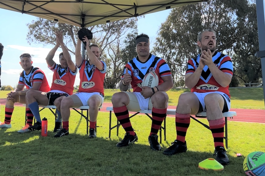 players wearing a red and sky blue rugby jersey with white shorts clap and cheer seated on a bench