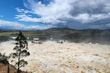 A river rages with heavy flows of brown water forming white wash, against a cloudy blue sky.