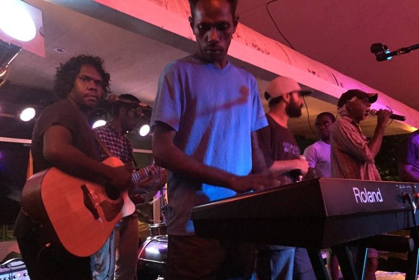 A group of young Aboriginal men playing instruments on stage and an old fella on the microphone.