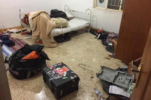 The room is messy with with supplies and camera equipment on the floor, and bed in the corner.