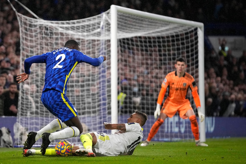A Leeds defender slides near the goal, taking the ball but also catching the legs of a Chelsea attacker in Premier League game.