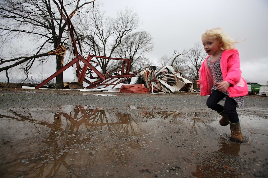 A girl wearing a pink jacket runs in front of a destroyed structure.