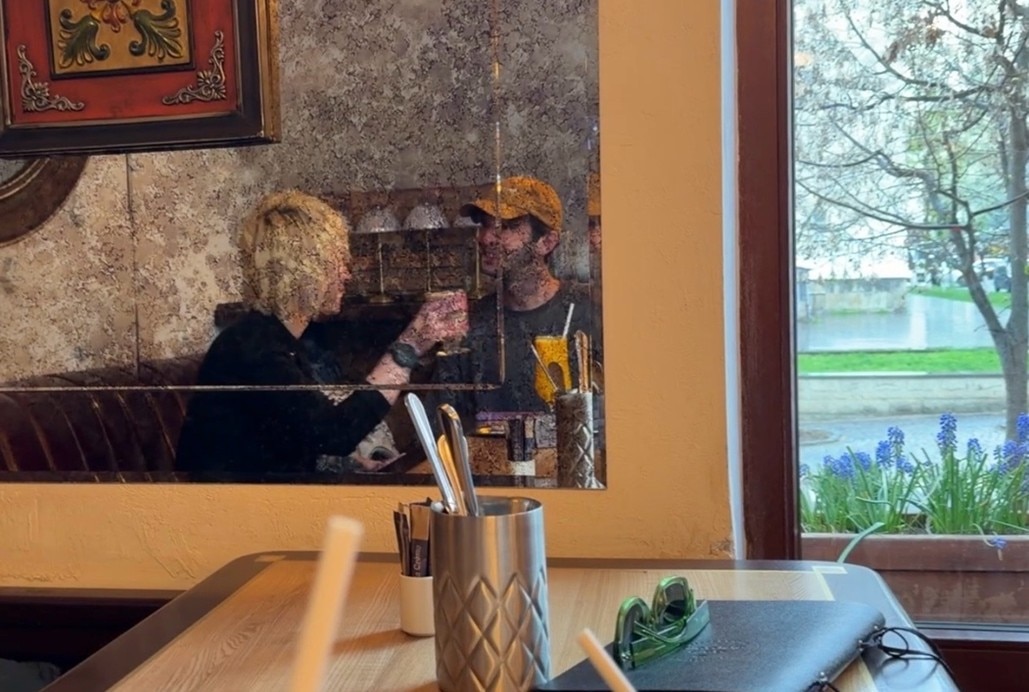 In the mirror, Elliot and Tatiana sit in a coffee shop chatting.