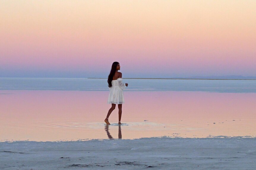 Une femme en robe blanche marchant sur un lac avec un coucher de soleil pastel en arrière-plan