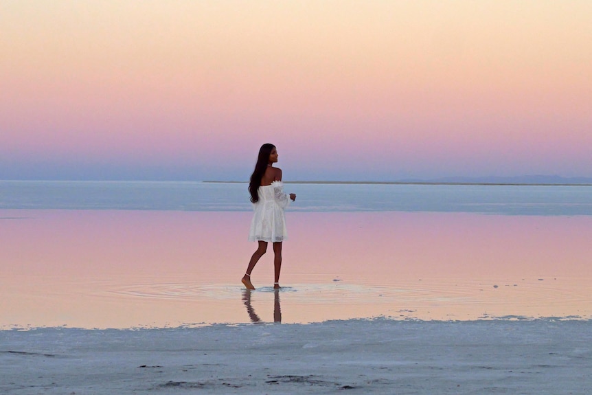 一个穿着白色连衣裙的女人在湖上行走，背景是柔和的日落