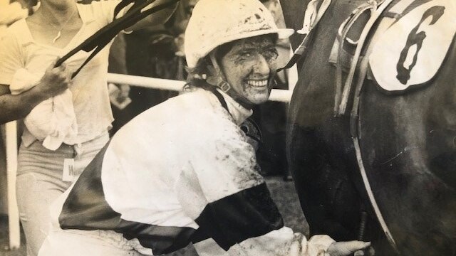 A woman in racing gear smiling and patting a racehorse