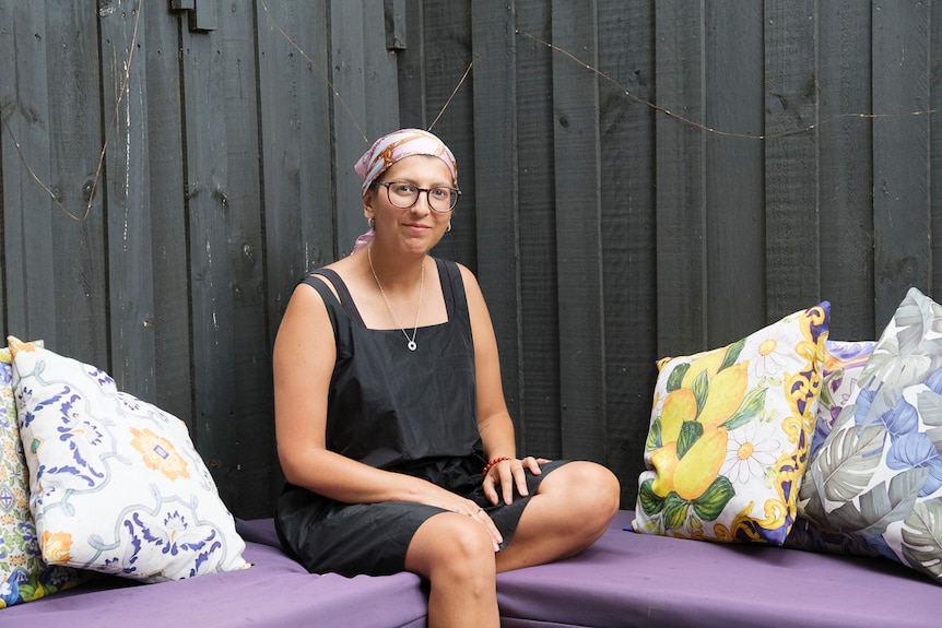 A woman wearing a black dress and a bright headscarf smiles at the camera. She is outside, surrounded by cushions.