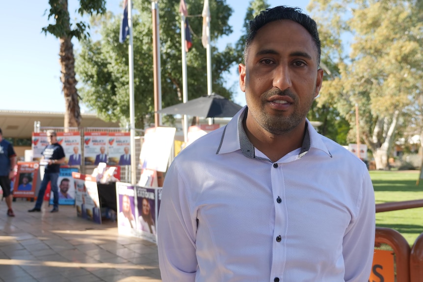 A man in a white collared shirt, with corflutes and election volunteers in the background.