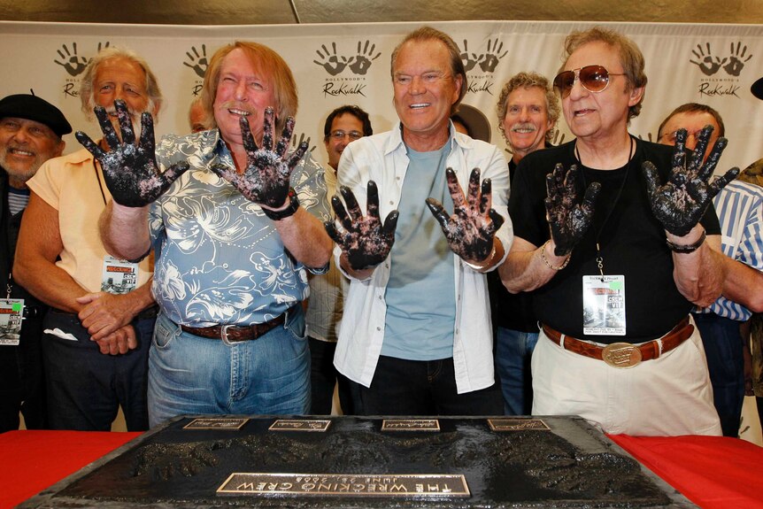 Don Randi, Glen Campbell and Hal Blaine, left to right, hold their hands up with wet cement on them. They are grinning.