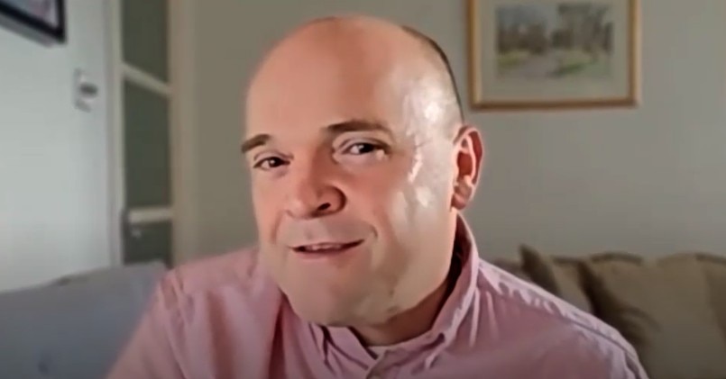 A man with a pink collared shirt smiles. Behind him is a framed picture on the wall.