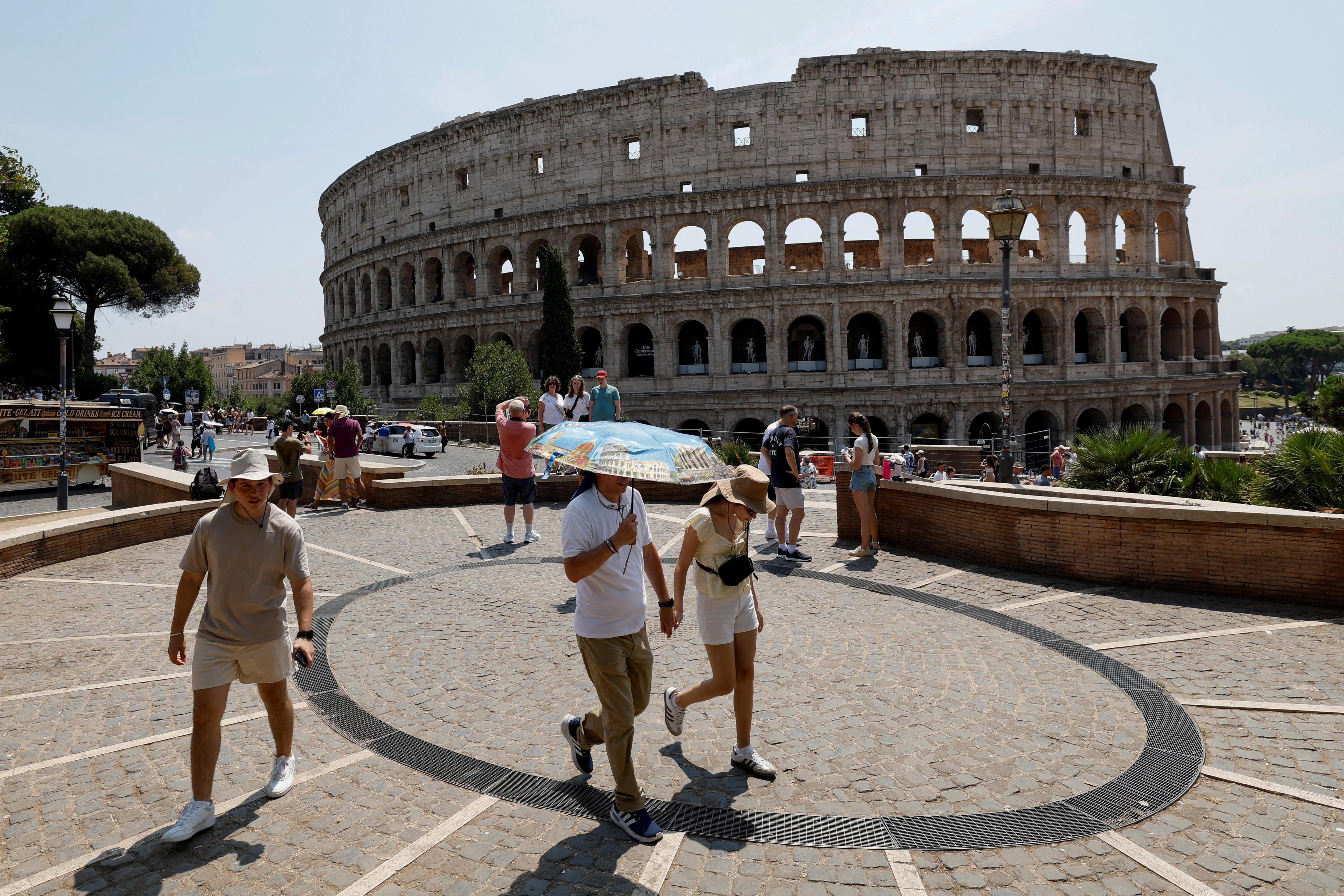 Pedestrians and tourists, including one under a blue umbrella, walking on a blue-sky day outside the Roman Colosseum.