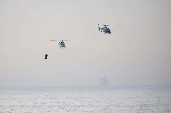 Franky Zapata is seen to the left as two helicopters fly above him. The sky is hazy and the sea is below. A ship is far behind.
