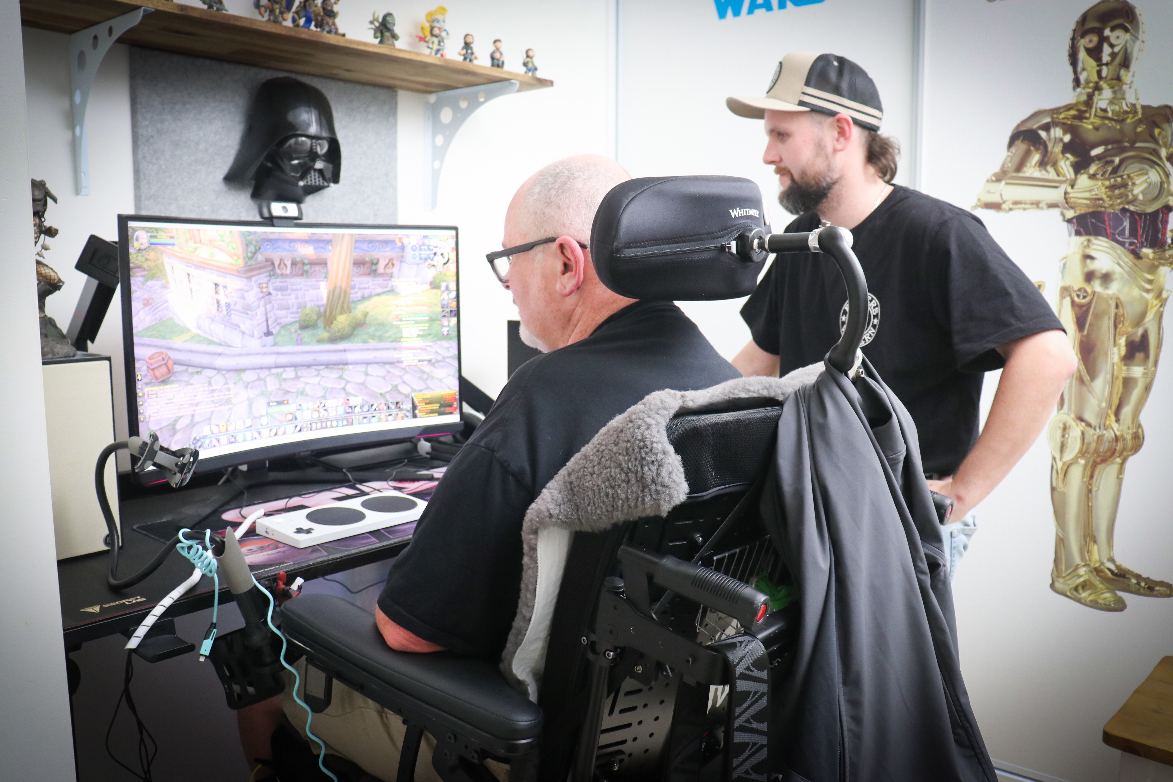 An older man in a wheelchair and a younger man with a beard watching a videogame on a computer screen.