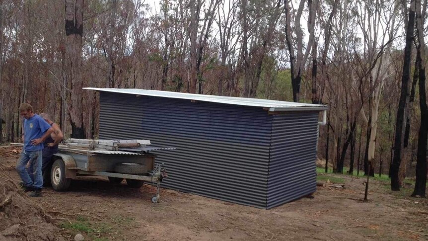 A shed with a flat white roof in a clearing in front of burnt-out trees.