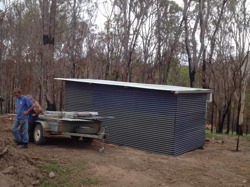 A shed with a flat white roof in a clearing in front of burnt-out trees.