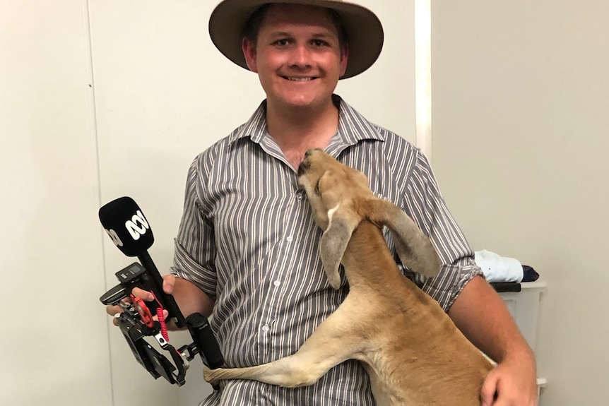 Man with hat and microphone cuddling a kangaroo.