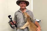Man with hat and microphone cuddling a kangaroo.