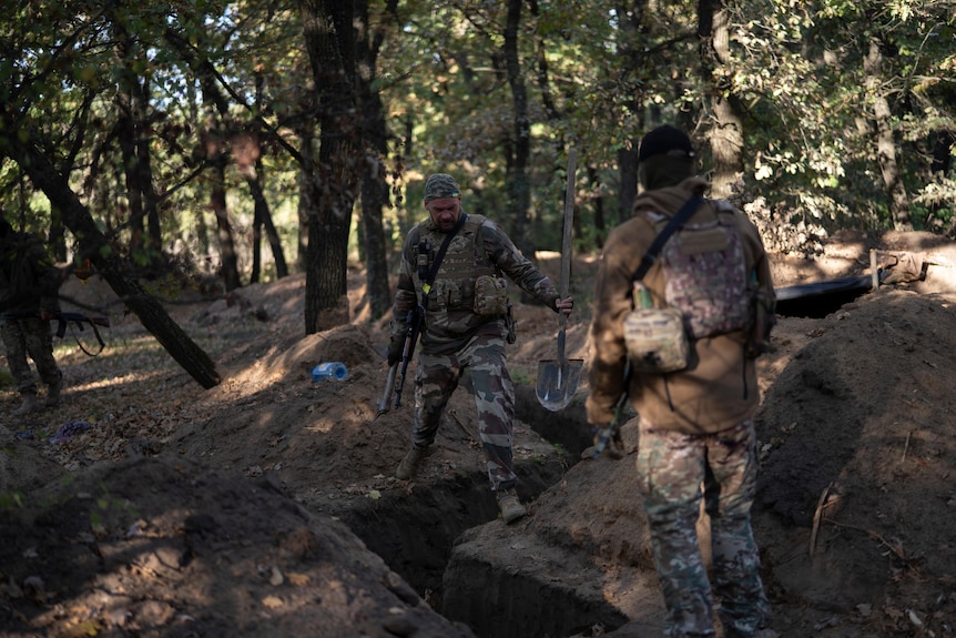 Two soldiers in combat gear look down into a trench in a forest.