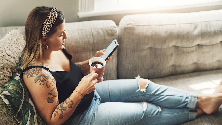 A woman wearing ripped jeans and black tank sits on a couch, looking at phone and drinking tea.