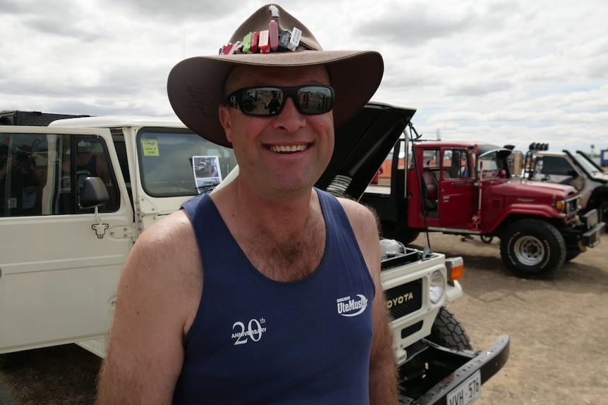 A man in sunglasses, a cowboy hat and a blue singlet smiles for the camera.