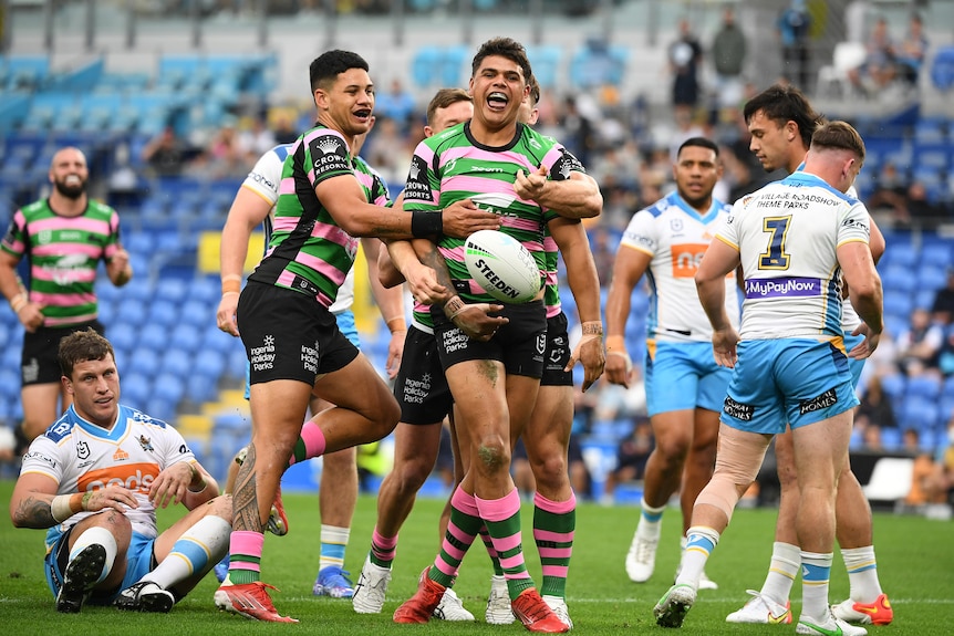 Latrell Mitchell grins and spins the football as his teammates grab in celebration after a try for Souths.
