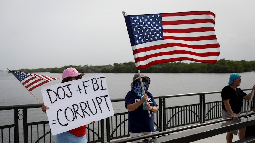 Two women holding American flags stand on a bridge in front of water. One is holding a sign that says "DOJ + FBI CORRUPT"