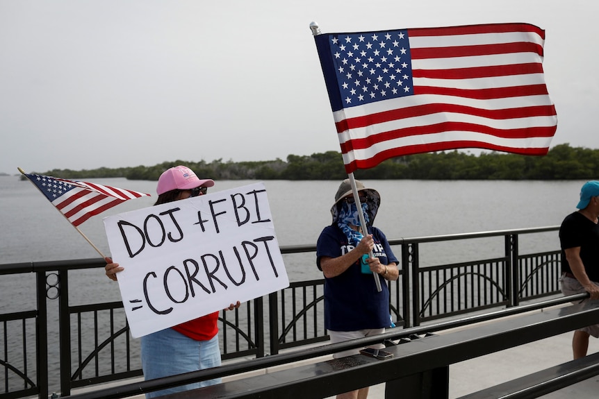 Two women holding American flags stand on a bridge in front of water. One is holding a sign that says "DOJ + FBI CORRUPT"
