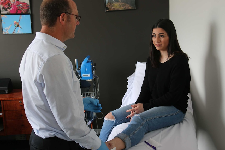 Wearing gloves, Daniel O'Brien looks at a wound on Jacinta Mazzarella's leg as she sits on a bed.