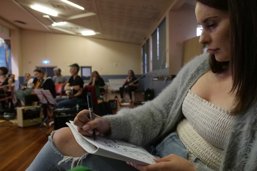 Young woman sketching on a notepad in the foreground, band members practicing behind.