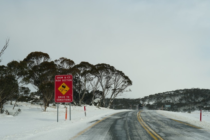 A snow and ice road sign on a landscape heavily blanked with snow, trees.