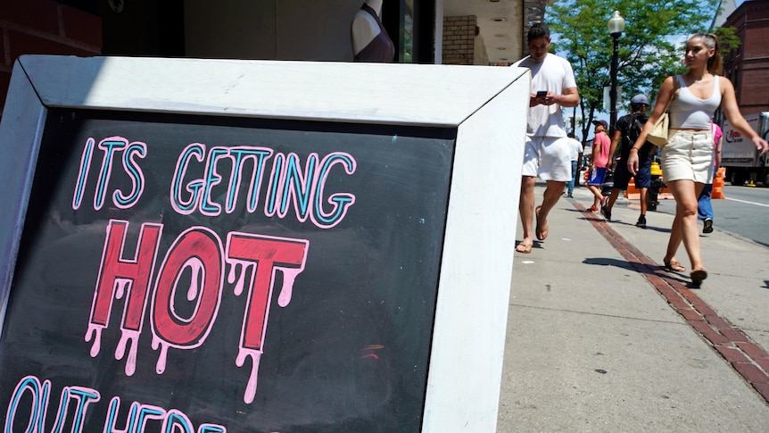 Pictured is a restaurant sign with the text 'it's getting hot out here', while people walk past it wearing summer outfits.