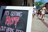 Pictured is a restaurant sign with the text 'it's getting hot out here', while people walk past it wearing summer outfits.