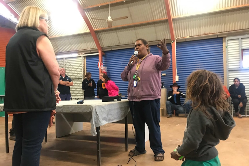 An aboriginal woman standing up speaking to a non-aboriginal health department worker in a large shed. People watch on.