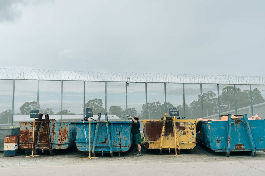 A prisoner puts metal inside a large metal bin with prison fence in background.
