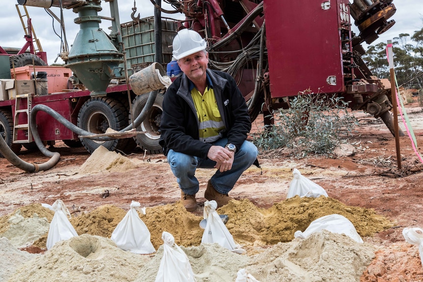 A man wearing a hard hat and high-vis workwear kneels down next to drilling samples in front of a drill rig.